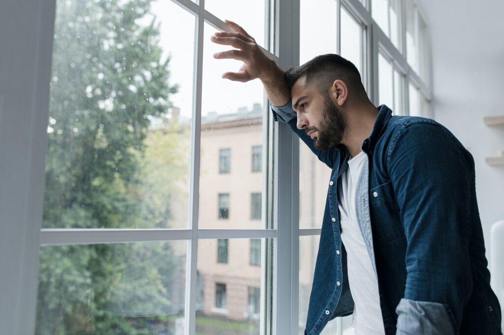 Sad young man with worried stressed face expression looking out window